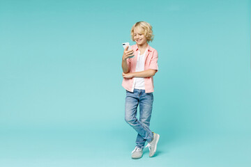 Full length of smiling little kid boy 10s in pink shirt using mobile cell phone typing sms message isolated on blue turquoise color background children studio portrait. Childhood lifestyle concept.