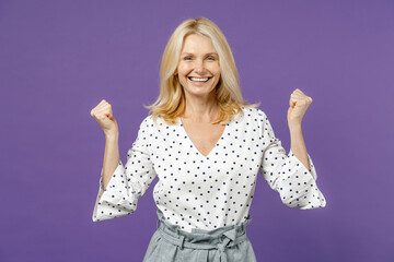 Wall Mural - Happy joyful elderly gray-haired blonde woman lady 40s 50s years old wearing white dotted blouse stand doing winner gesture clenching fists isolated on bright violet color background studio portrait.