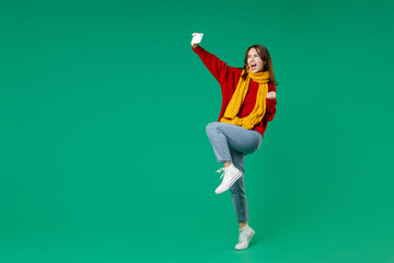 Wall Mural - Full length side view of happy young brunette woman 20s in knitted red sweater yellow scarf doing selfie shot on mobile phone doing winner gesture isolated on green color background studio portrait.