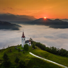 Wall Mural - Šebrelje, Slovenia - Aerial drone view of the beautiful hilltop church of St.Ivan (Sv. Ivan Cerkev) at sunrise with huge morning fog bellow the valley and Julian Alps at background