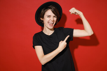 Wall Mural - Laughing strong sporty young brunette woman 20s in basic black t-shirt hat standing point index finger on biceps muscles on hand looking camera isolated on bright red color background studio portrait.