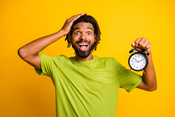 Wall Mural - Photo of young man amazed shocked hand touch head hold alarm clock late overtime wear green t-shirt isolated over yellow color background