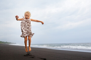 Happy family have fun on tropical sea beach resort. Funny young girl jumping high at black sand beach. Active lifestyle, people travel activity on summer holiday with kids