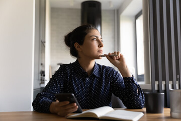 Canvas Print - Pensive young Indian woman write in notebook use cellphone look in distance pondering thinking. Thoughtful millennial female plan decide make notes working on smartphone at home office.