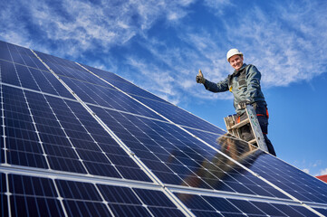 Wall Mural - Low angle view portrait of a smiling worker, installing solar batteries, who is standing on ladder at solar plant against blue sky, showing thumb up. Concept of alternative sources of energy.