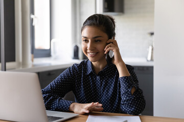 Canvas Print - Smiling millennial Indian woman work on laptop from home office talk on cellphone consulting client or customer online. Happy young ethnic businesswoman have smartphone consultation with partner.