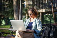 A Young Serious Woman Student Sitting On A Bench In The Park And Typing On Her Laptop On A Sunny Day In Universiry Campus