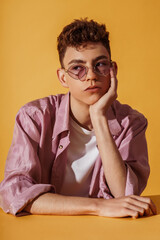 Close up studio fashion portrait of young man wearing trendy sunglasses, silk lilac color shirt, white t-shirt, posing on yellow background
