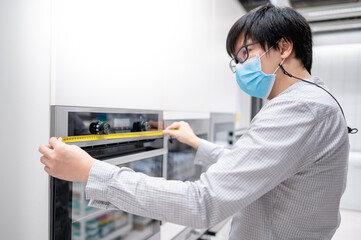 Wall Mural - Asian man interior designer wearing face mask using tape measure on oven in kitchen showroom at furniture store. Handy cooking appliance for domestic kitchen. Home improvement concept