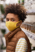 Vertical Portrait Of Teenage African-American Boy Wearing Mask In Supermarket And Looking At Camera With Shelves In Background