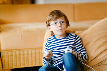 Cute little blond kid boy with glasses playing with a video game console. Child having fun at home during corona virus lockdown quarantine. Lonely alone boy without friends, indoors