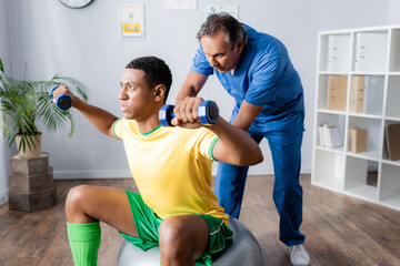 african american man in sportswear training with dumbbells on fitness ball near physiotherapist