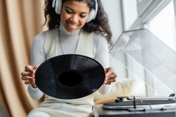 happy african american woman in wireless headphones holding vinyl disc near record player, blurred foreground