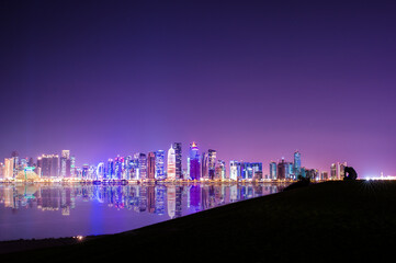 (Selective focus) Stunning panoramic view of the Doha skyline illuminated at dusk in the distance and some people relaxing on a hill in the foreground.