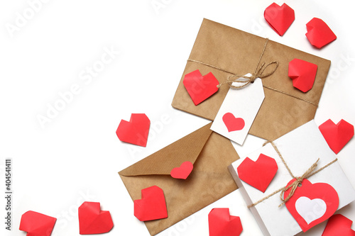 White gift box with red label in a heart form, gift wrapped in brown craft paper, tied with twine with a bow, envelope from craft paper with red heart, several handmade red 3D paper hearts on white 