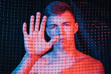 Prisoner loneliness. Defocused portrait. Human trafficking. Freedom protection. Hopeless shirtless man trapped behind mesh fence in red blue neon light on blur dark background out of focus.