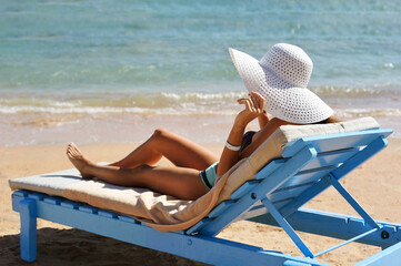 Wall Mural - Beautiful woman sunbathing on a beach at tropical travel resort, enjoying summer holidays. Girl holding a hat with her hands on sun lounger near sea