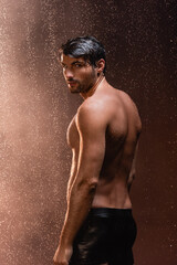 sexy man with muscular torso looking at camera while posing under rain on dark background