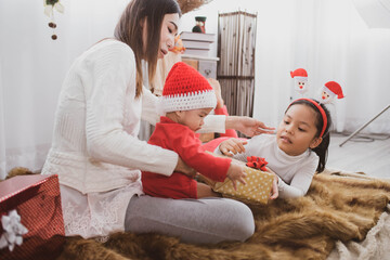 Wall Mural - parent and two little children having fun and playing together near christmas tree indoors. merry christmas and happy holidays. cheerful mom and her cute daughters girls exchanging gifts.