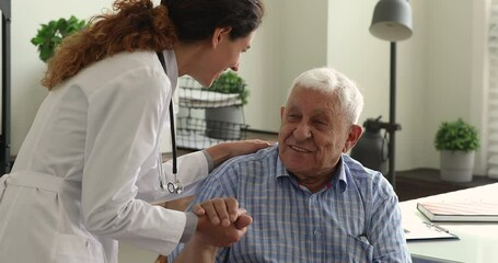 Wall Mural - Young gp doctor holds senior male patient hand talk to him provide psychological support express kindness, give professional aid during visit in clinic. Elder care, medicare services for older concept