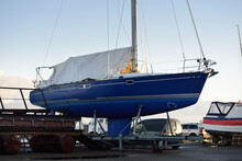 Blue Sloop Rigged Yacht Standing On Land In A Small Harbor On A Clear Winter Day. Waiting For The New Sailing Season. Industry, Business, Transportation, Service, Logistics, Sport, Recreation Concepts