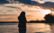 © shchus - Silhouette of a young beautiful caucasian girl in jeans and a jacket against the backdrop of a cloudy sunset and water, sea or river. The concept of loneliness and self-sufficiency.