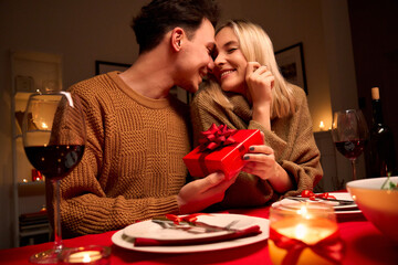 Happy young couple celebrating anniversary or Valentines day having romantic dinner at home table. Loving man giving red gift box hugging beloved woman making present surprise on date in candle light.