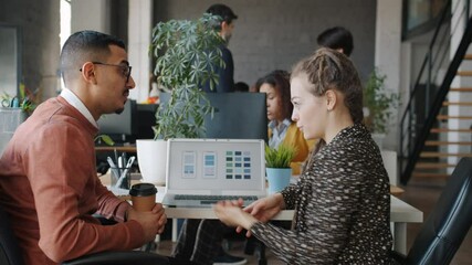 Arab man and Caucasian woman are discussing smartphone application development using computer talking looking at screen sitting at desk in shared office