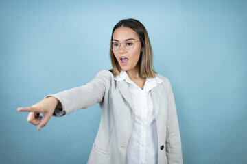 Young business woman over isolated blue background pointing with finger surprised ahead, open mouth amazed expression, something on the front