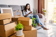 © Kittiphan - Happy young asian woman using smartphone in living room at new house with stack of cardboard boxes on moving day