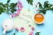 Paper sheet with text HELLO SPRING, tea and floral decor on color background