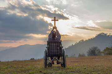 Wall Mural - Paralyzed woman sitting on her wheelchair and hold the crucifix in hands while praying to God at sunset background