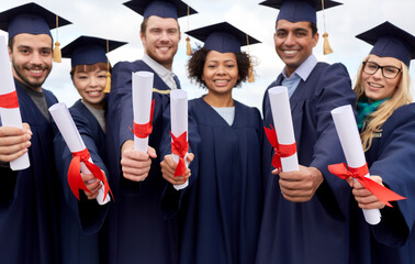 education, graduation and people concept - group of happy international graduate students in mortar boards and bachelor gowns with diplomas