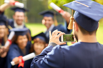 Poster - education, graduation, technology and people concept - group of happy international graduate students in mortar boards and bachelor gowns with diplomas taking picture with smartphone outdoors