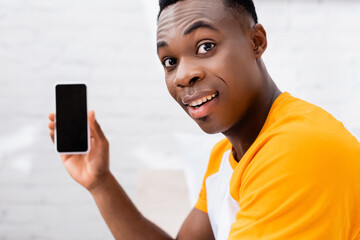 Smiling african american man looking at camera while holding smartphone with blank screen on blurred background