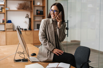 Wall Mural - xSmiling woman talking on cellphone and working with computer in office