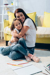 Cheerful mother with closed eyes hugging daughter while sitting on floor on blurred background