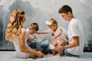 Smiling mom and dad with daughter, son sitting on the floor. Fashion models plays with feathers. Happy family portrait in casual style clothes. Nice family wearing jeans isolated on grey wall.