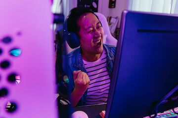 Young Asian male gamer won and victory the winning game with headset and pc computer. Overjoyed live streamer streaming video game in low light room. Excited gaming influencer smiling. With copy space