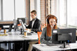 © Roman - woman manager takes cold calls on the phone, talks with clients sitting in the office with colleagues, using headphones