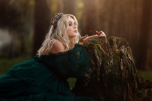 A Beautiful Blonde Young Woman In A Long Green Dress And A Diadem On Her Head In The Forest. Girl Sitting Near The Old Stump With Butterfly On Her Hand. Solar Glare. Fantasy. Fairy Tale
