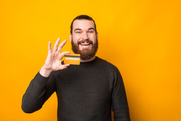 Wall Mural - Cheerful bearded man smiling and showing credit card over yellow background.