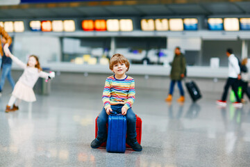 Cute little kid boy with luggage, two suitcases on international airport. Mother with little girl daughter on background, Happy family wating for flight and going on vacations. Travel lifestyle.