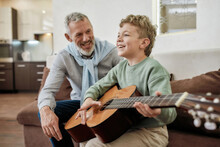 Excited Little Boy Learning How To Play Acoustic Guitar With Grandfather, They Sitting Together On Sofa In The Living Room And Laughing, Having Fun
