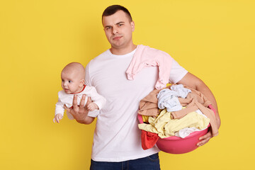 Single father overwhelmed by taking care of everything alone, take care of new born baby and doing laundry, wearing casually, stands isolated over yellow background, holds basin with clean clothing.