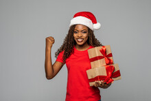 African Woman In Santa Hat With Win Gesture Christmas Mood Carrying A Gift Box Isolated On White Background