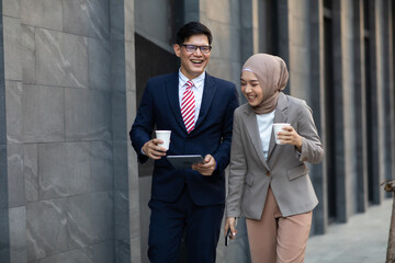 Wall Mural - Young business muslim woman and business man colleague walking and discussing outside the office. Woman is holding a coffee and businessman is holding a digital tablet outdoor.