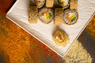 Top view of Japanese Tempura hot sushi roll with roasted salmon served with wasabi, ginger on white square plate. Dark background with scattered spices: dried garlic, turmeric, curry, paprika powder
