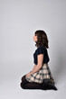 Full length portrait of pretty brunette woman wearing tartan skirt and blouse.  Sitting pose on the ground, against a  studio background.