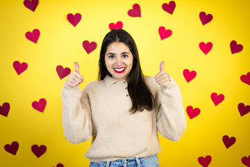 Young caucasian woman over yellow background with red hearts success sign doing positive gesture with hand, thumbs up smiling and happy. cheerful expression and winner gesture.